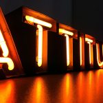 Neonletters vormen het woord "ATTITUDE" in fel oranje, met reflecties op een glanzend oppervlak.