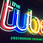 Felgekleurde neonletters vormen de tekst "the tube" en "UNDERGROUND EXPERIENCE" op een donkere achtergrond.