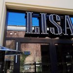 Verlichte letters "LISA" met spots, boven een glazen ingang en een metalen terrasverwarmer op de voorgrond.