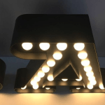 Zwart metalen letter met ronde lampjes erin, die samen een opvallende vorm creëren op een lichte achtergrond.