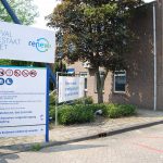 Een grijze kantoorgebouw met een blauwe en witte bord aan de ingang, omgeven door bomen en parkeerplaatsen.