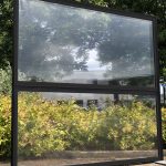 Een groot, zwart frame met een doorzichtig paneel, omringd door groene struiken en bomen in de achtergrond.