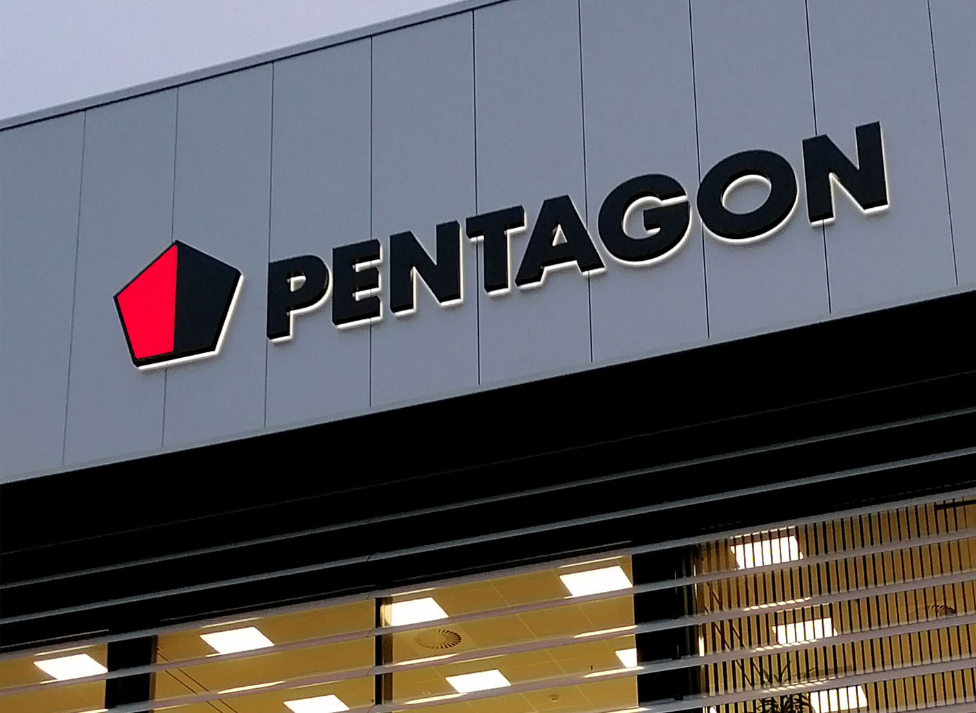 Logo van Pentagon op een gevel met een rode vijfhoek en zwarte letters, verlicht tegen een grijze achtergrond.